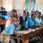 Seven young people wearing glasses sit in the front row on a wooden school bench. They are all wearing blue shirts and looking into the camera.