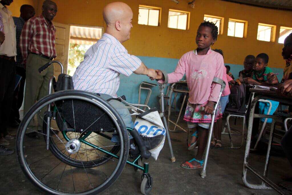 White man in wheelchair shakes hand with young girl from Mozambique who uses crutches. They are in a brightly coloured yellow and blue school classroom.