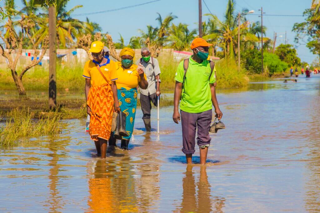 4 Mozambican adults wade through a flooded street. At the rear is an older man with one leg and crutches.