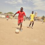 Two members of the Sports for Peace team, one in red and one in yellow, play football on a dusty pitch in South Sudan.