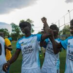 Four players from the South Sudan blind football league. Two players are holding aloft the arm of another player. The three are smiling as they come off the pitch and wearing football shirts with Blind Football on the front.