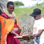 Kushuke Legei with David Ndungu, Disability Inclusion Field Officer at Light for the World. Kushuke is wearing an organge shawl with colourful clothes and jewellery. She is looking at a training manual with David, who points something out on a page. They are in Kushuke's home village.