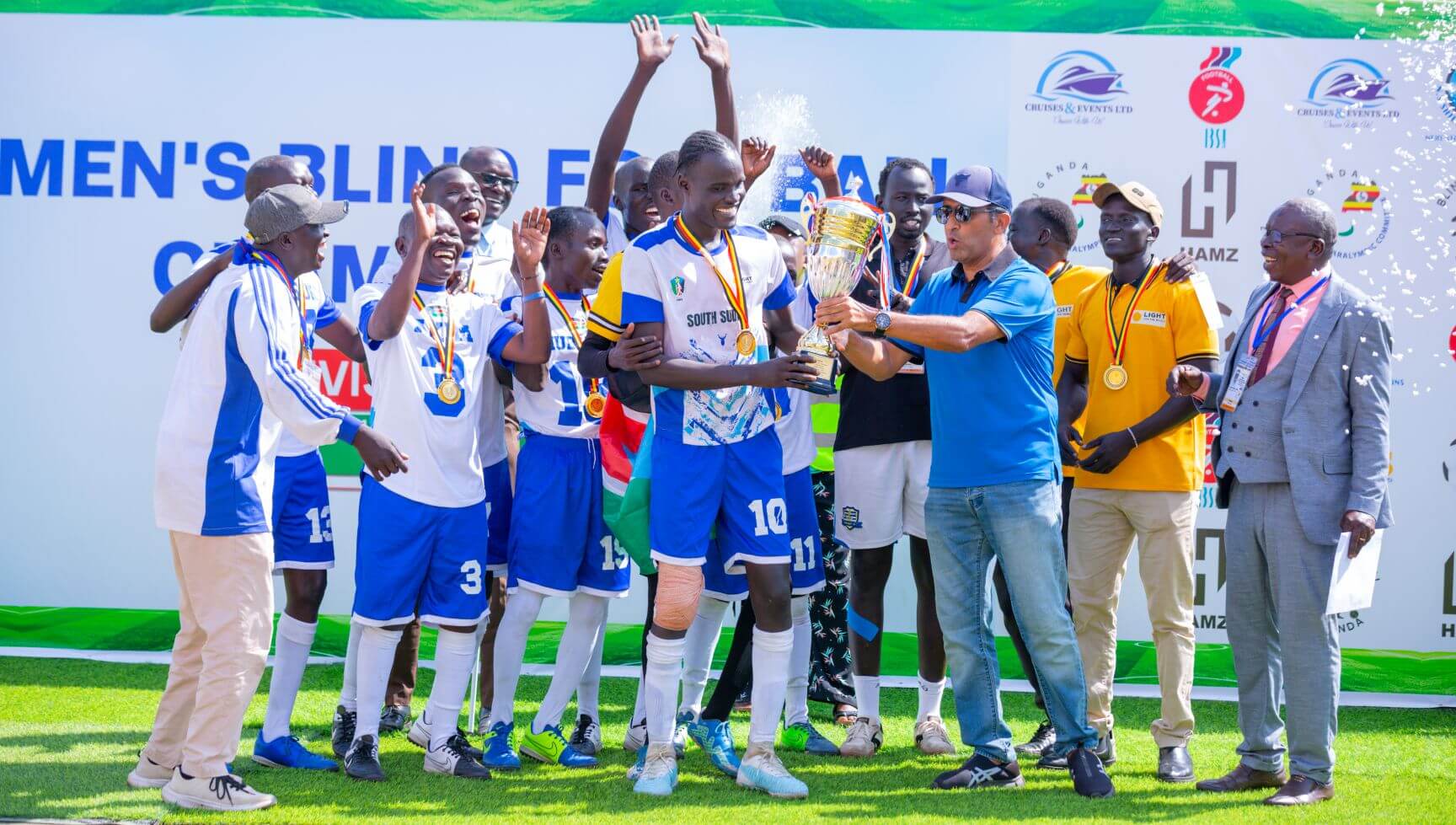 South Sudan players in white and blue kits celebrate as captain Martin Ladu Paul is presented with the championship trophy. Medals hang around their necks, players hold their hands aloft in celebration and everyone is smiling.