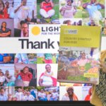 A woman smiles and holds up a copy of the Light for the World Kenya Country Strategy 2026–2030. Behind her is a large screen displaying a photo collage of people supported by Light for the World Kenya's programmes, with the Light for the World logo and the word 