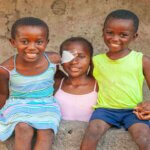Three smiling children sit together on a concrete step in Quelimane, Mozambique. In the centre is Fatima, who wears a pink top and a bandage over her eye after cataract surgery. She is flanked by a girl in a striped blue dress and a boy in a bright yellow top.