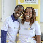 Deborah Achieng (left) and Maureen Thuku (right), both participants from Zetech University, stand together smiling and wearing white T-shirts co-branded with the Standard Chartered Foundation and Light for the World logos. A Light for the World Kenya banner is visible behind them.