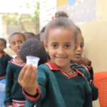 Aryam Teklay, a young girl in a school uniform, holds up a small cup of medication during a trachoma treatment campaign, with classmates lined up behind her.