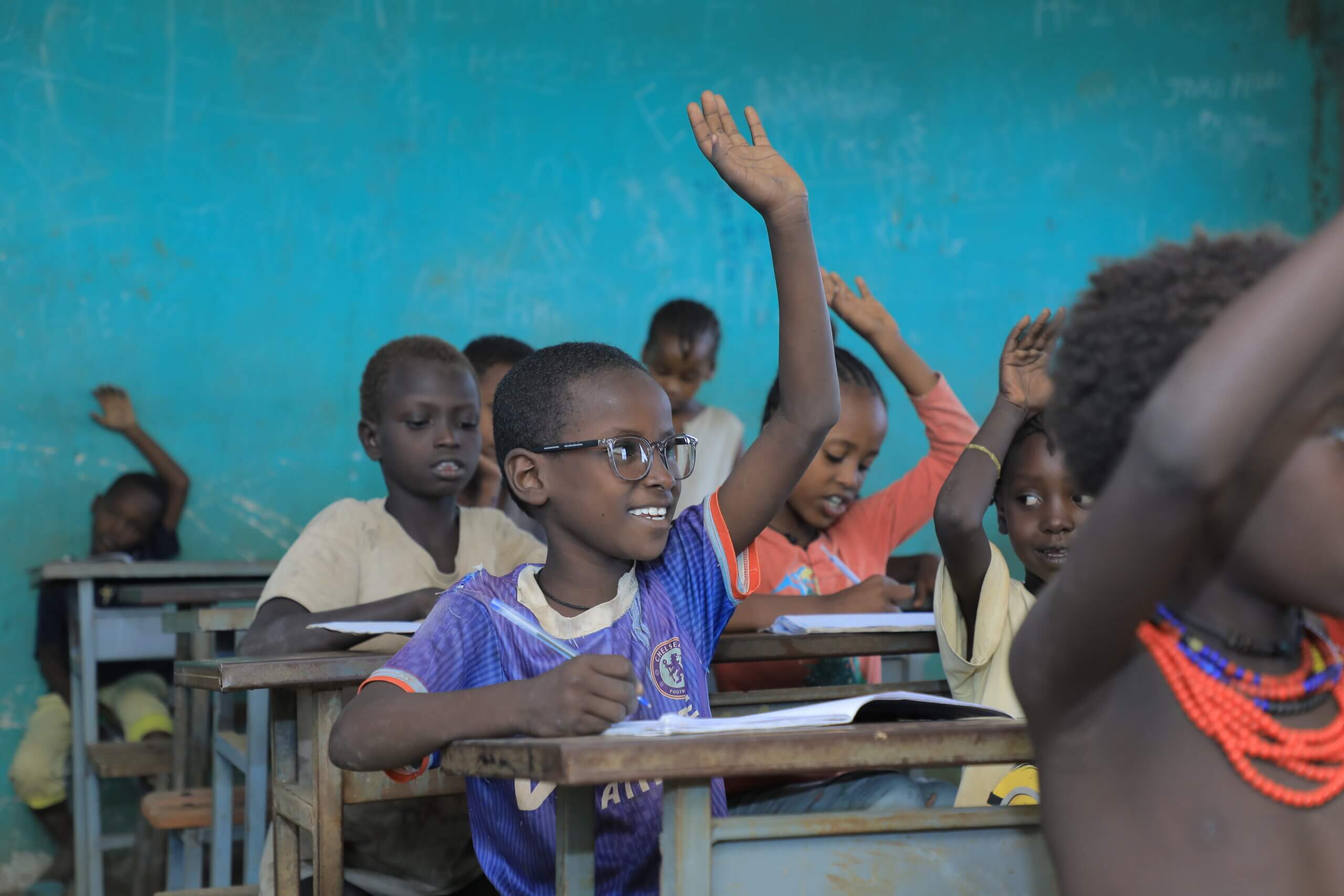 A classroom of children. The image focuses on one boy, wearing a blue sports t-shirt and glasses with clear frames. He has a smile on his face, and holds a pen in one hand, and the other hand up in the air. The classroom has blue walls and wooden desks, and we can see seven other children sitting around him, some also with their hands in the air.