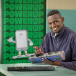 Benson fixes a power socket extension. He is sitting at a green table, holding the extension in his left hand, the other hand holding a small screwdriver. He wears a navy fleece, has short dark hair, and smiles at the camera. There is a keyboard on the desk in front of him, and behind a green banner reads “PowerUp” and shows a graphic illustration of a smiling mobile phone with arms and legs.
