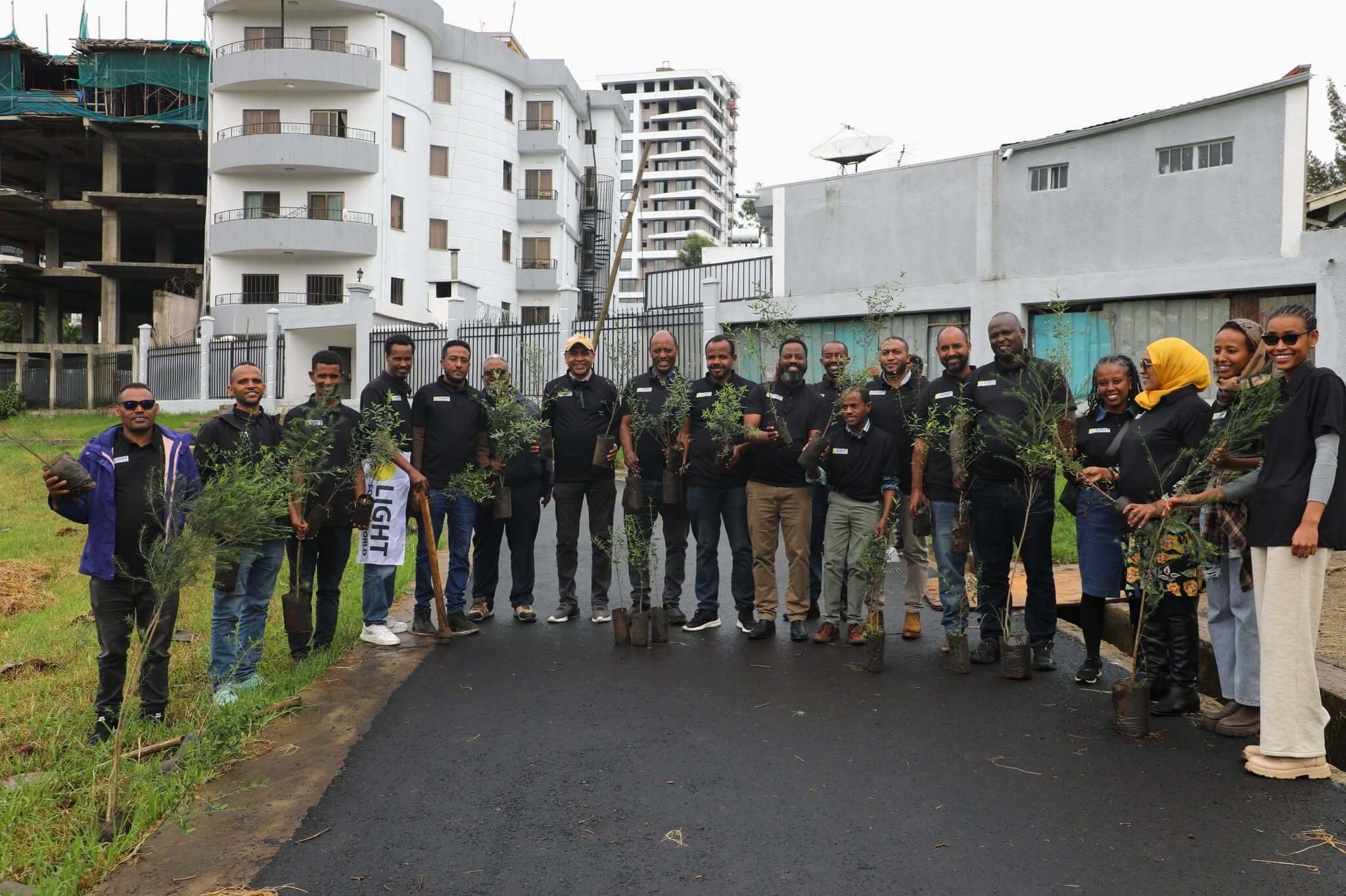 The Light for the World Ethiopia team stand outside, holding small green trees, to be planted. They are all wearing black t-shirts with the “Light for the World” logo on the chest. There is a white building and some construction in the background, with a green grassy area to the left. It looks like it has been raining, with a few people wearing hats, headscarves and coats.