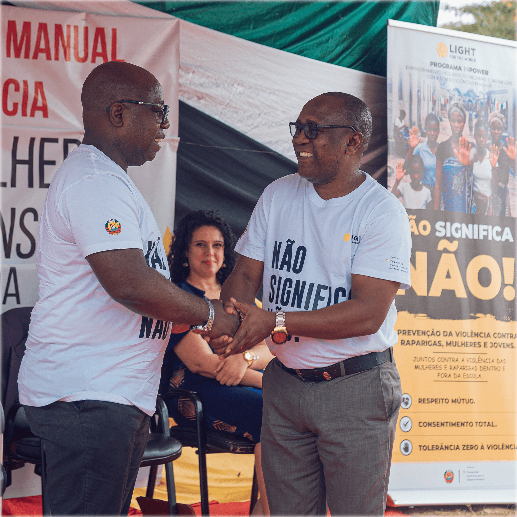 Two men, both wearing glasses and white t-shirts, which read “não significa não” (no means no), shake hands. They seem to be standing on a stage, and are in front of two banners, you can see the word “manual” in red on one and “não significa não” on the other. A woman sits on a black chair, smiling in the background.