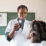 Teresa Tomas Mussa, a teacher trained in pre-screening eyes and wearing a white coat, shines a light from a smartphone into a pupil's eye during a classroom vision check. An eye test chart is pinned to the chalkboard behind them.