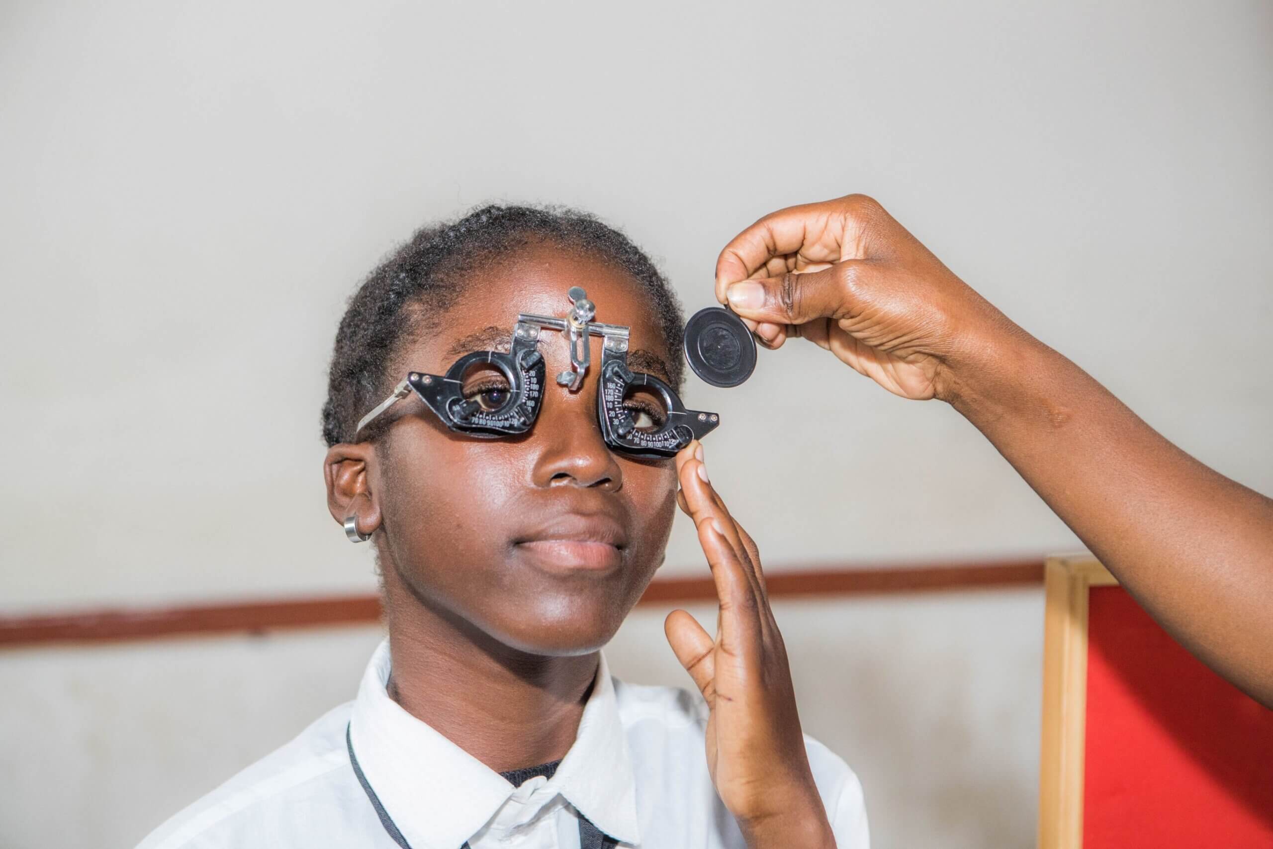 A girl wears a metal trial frame with an occluder over one eye while a technician holds a trial lens up to test her vision, in a room with a red support pillar.