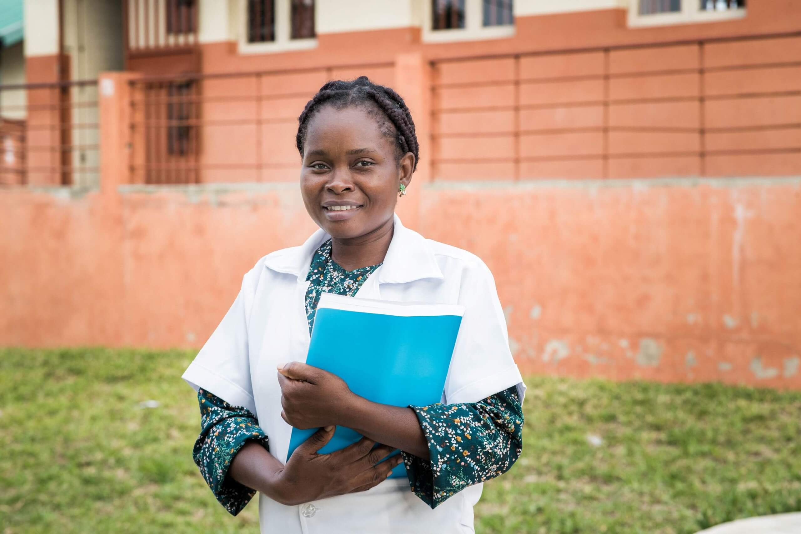 Teresa Tomas Mussa, wearing a white coat over a green floral top, stands smiling outdoors holding a folder of school papers, in front of a brick perimeter wall.