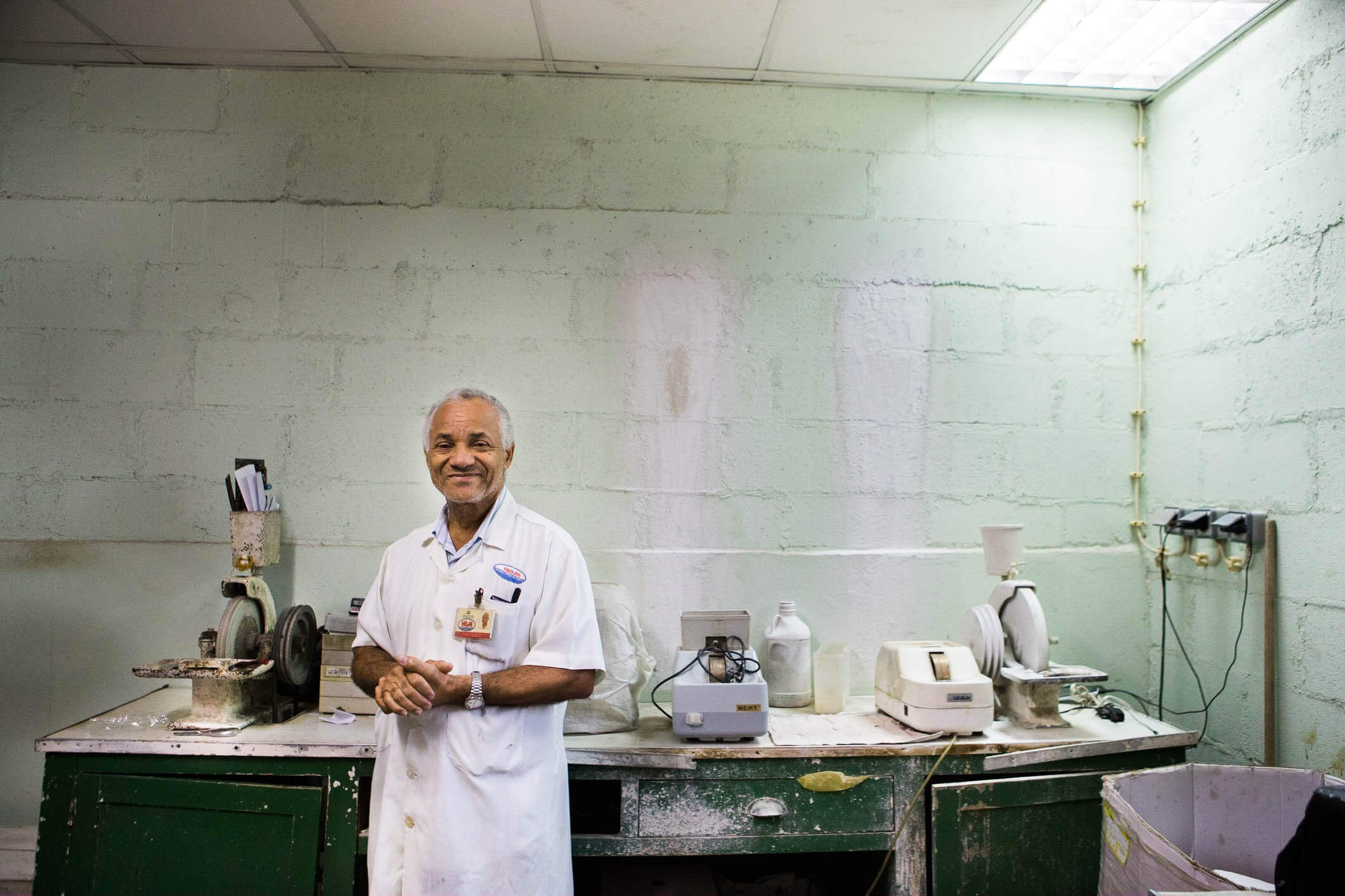 Neutel Simoes de Abreu, an older man in a white coat with an "HGM" name badge, stands smiling with hands clasped in a worn workshop with mint-green painted brick walls. Old grinding and polishing machines, bottles, and hand tools sit on a green wooden bench beside him.