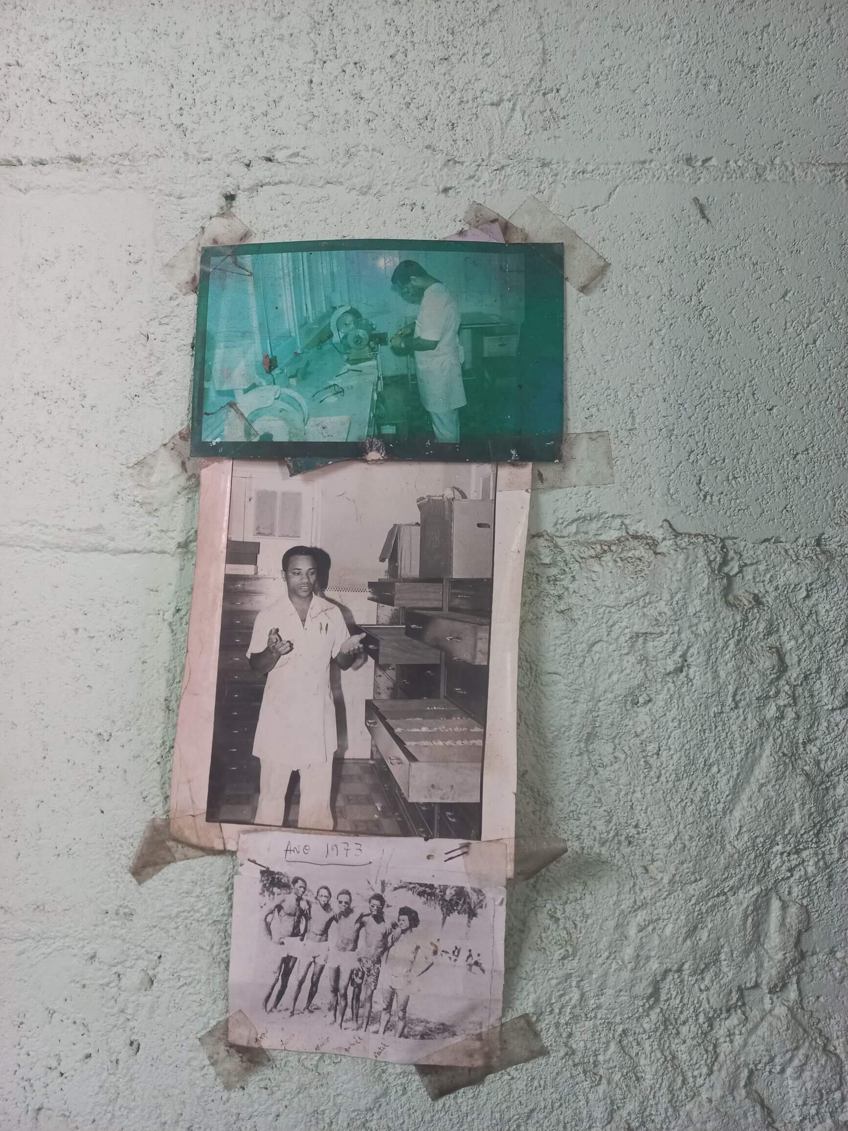 Three old photographs taped to a rough painted wall. The top photo, tinted teal with age, shows a man working at a grinding machine. Below it, a black-and-white photo shows a man in a white coat standing among open drawers of equipment. The bottom photo, labelled "Ano 1973" by hand, shows a group of young men posing together outdoors.
