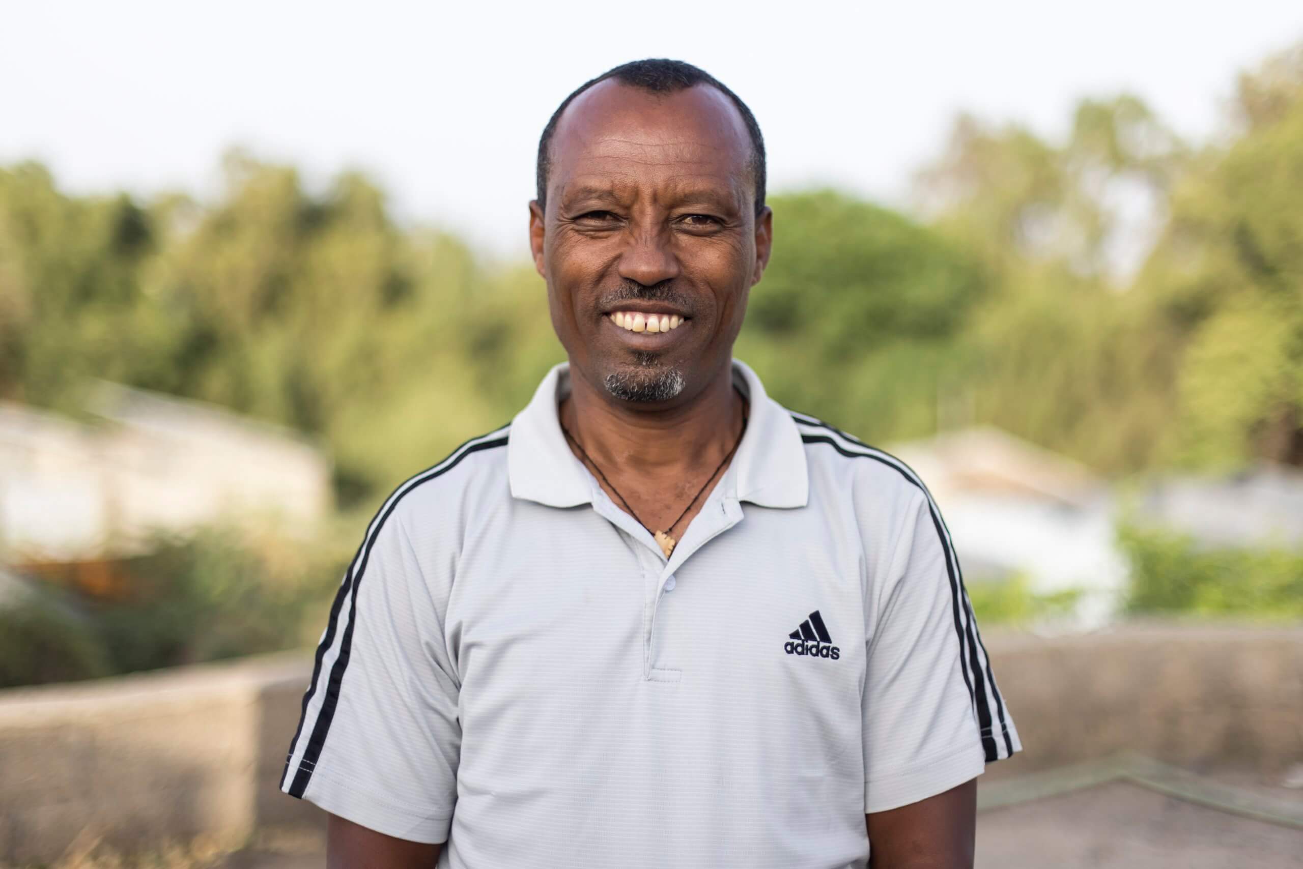 Tefera Hiluf smiles and looks at the camera. He has short dark hair, a moustache and a small beard on his chin. He is standing up outside, wearing a white adidas polo shirt, with black stripes, and a necklace tucked underneath. The background is out of focus, with blurry green trees and white buildings.