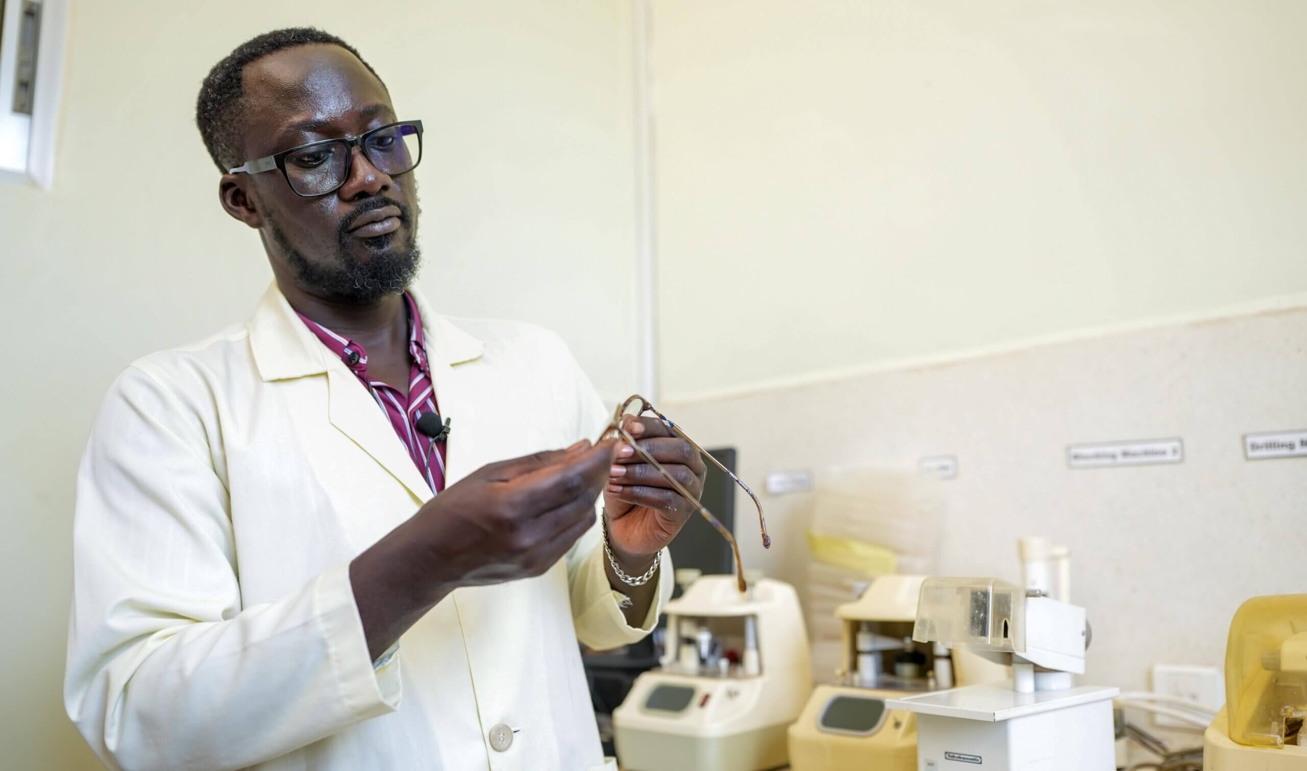 Jimmy Ogwal, wearing a cream lab coat and glasses, closely examines a pair of spectacle frames held in both hands. Workshop machinery is visible on the counter behind him.