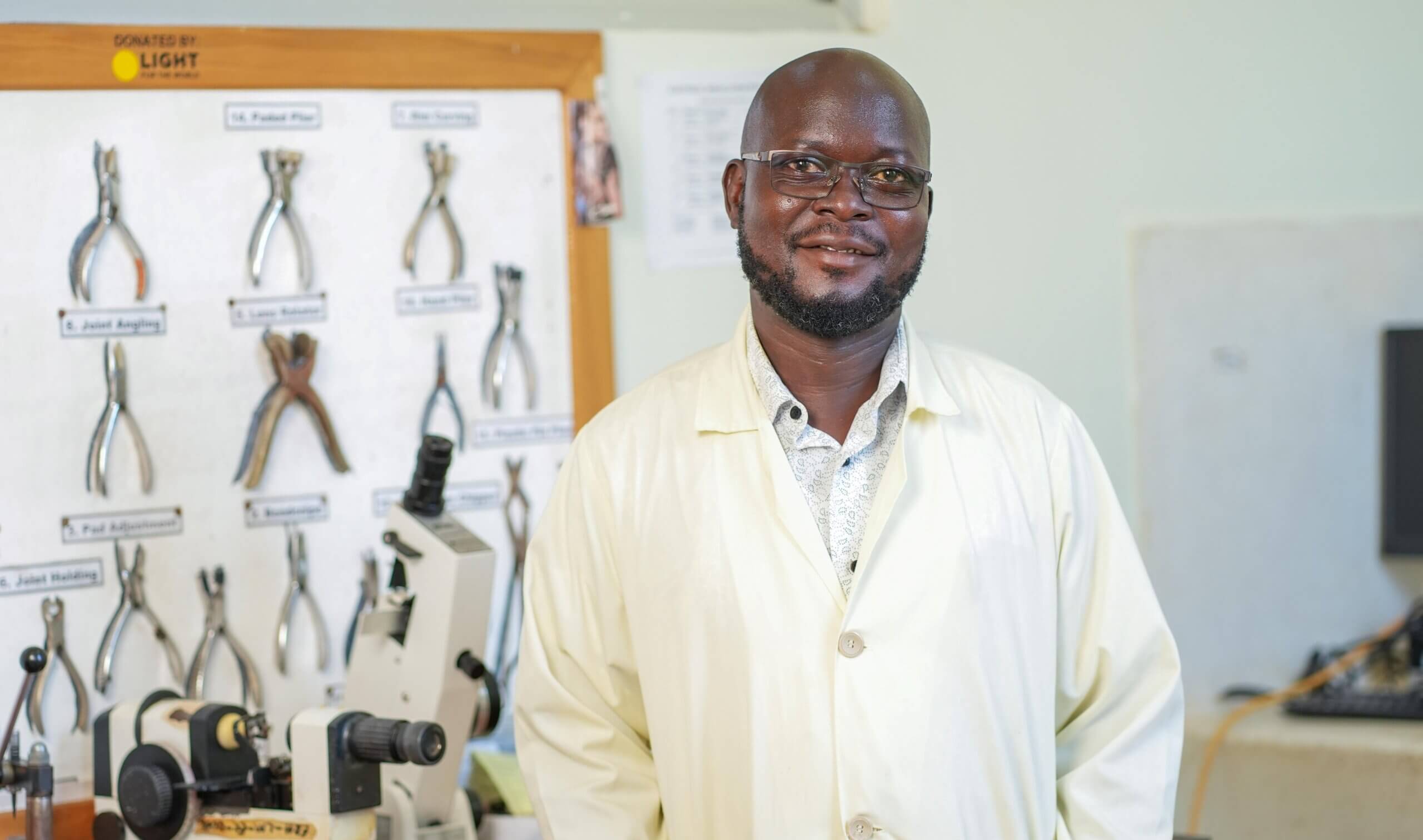Davidson Komakech, wearing glasses and a cream lab coat, stands smiling in front of a board displaying labelled pliers and workshop tools, next to a lensometer.