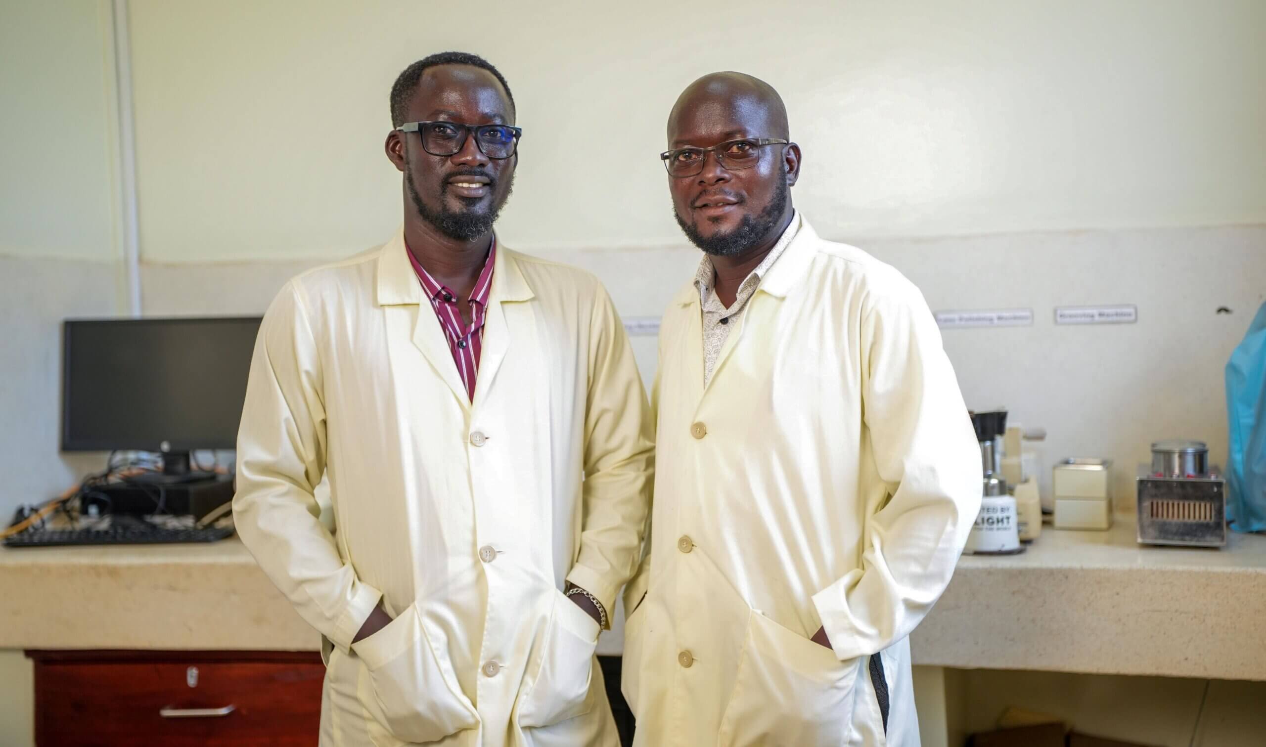 Jimmy Ogwal (left, wearing dark-framed glasses) and Davidson Komakech (right, bald) stand side by side in cream-coloured lab coats in an optical workshop, smiling at the camera. Behind them, a counter holds equipment, cables, and a monitor.