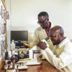 Davidson Komakech, seated, looks through a lensometer to check a lens, while Jimmy Ogwal stands beside him holding a slip of paper and watching closely. Optical tools hang on a board to the left.