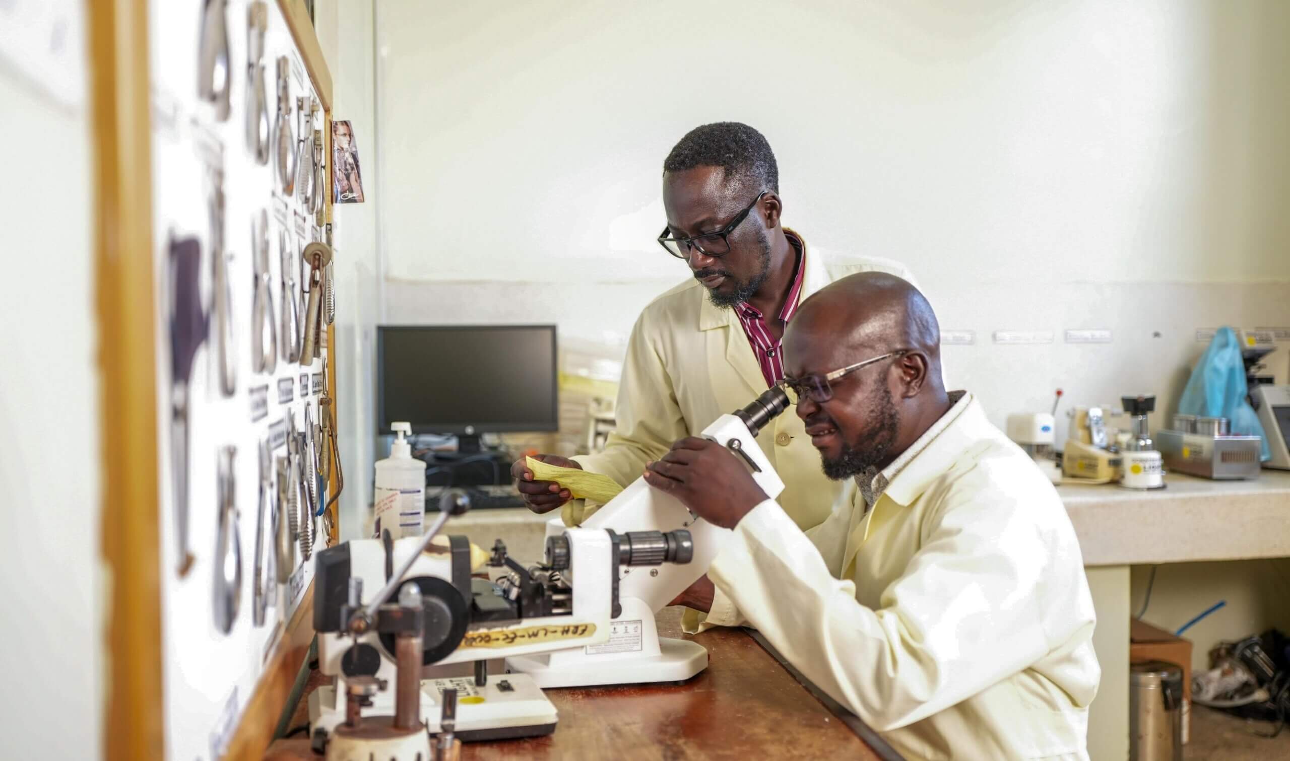 Davidson Komakech, seated, looks through a lensometer to check a lens, while Jimmy Ogwal stands beside him holding a slip of paper and watching closely. Optical tools hang on a board to the left.