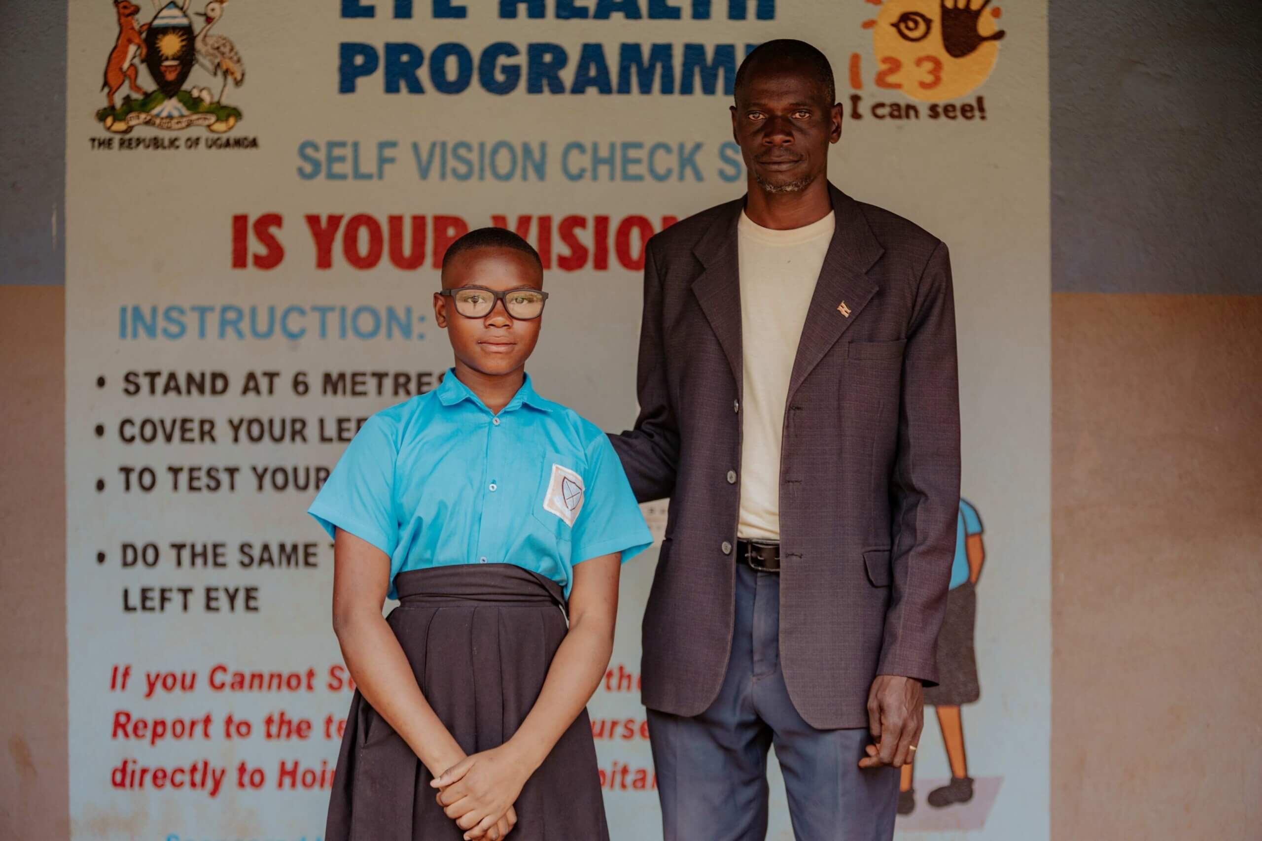Godfrey stands with pupil Cleopatra, who wears glasses and a blue school uniform, in front of a "vision wall", which allows students to self-check their vision.