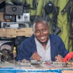 Aaron Masia, a man with a short beard, wearing a navy blue coat over a striped shirt, smiles at the camera while working on a circuit board on a cluttered workbench, surrounded by shelves of electronics and hanging cables.
