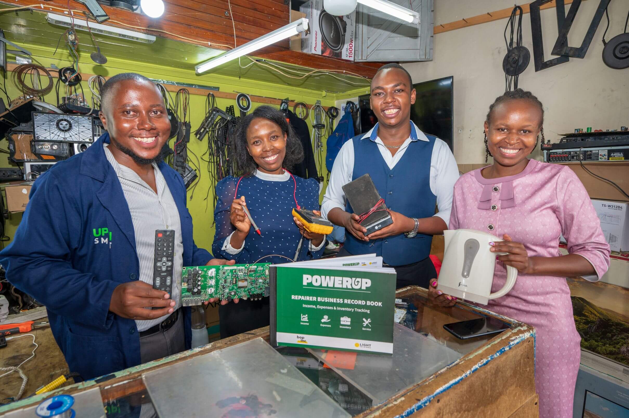 Four smiling people stand behind a glass display counter, each holding a repaired item — a TV remote, a green circuit board, a multimeter, a battery pack, and a white electric kettle. A green "PowerUp Repairer Business Record Book" sits on the counter in front of them, in a workshop lined with wires and speakers.