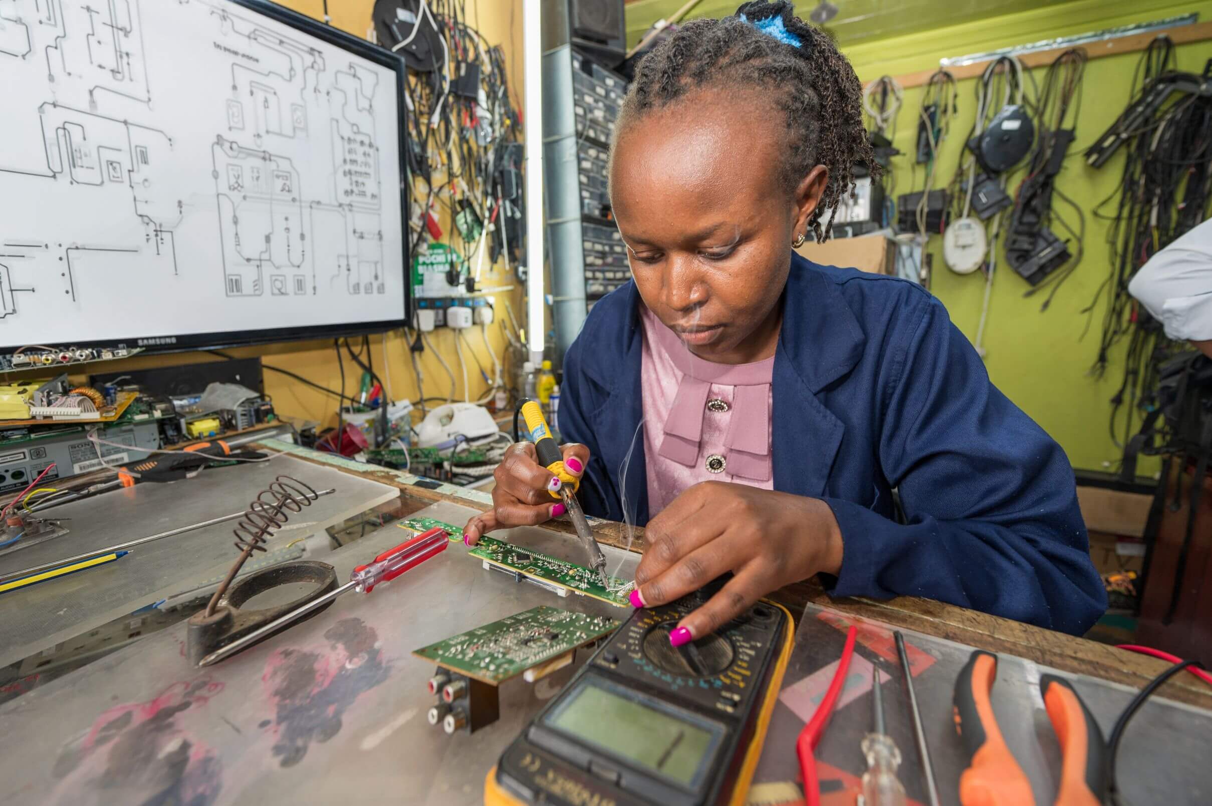 Jane Waweru, a woman wearing a navy blue coat over a pink top, solders a circuit board at a workbench, a soldering iron in one hand and a multimeter in the other, with smoke rising from the joint. A wiring diagram is displayed on a repurposed TV screen in the background, surrounded by cables and spare parts on a green wall.