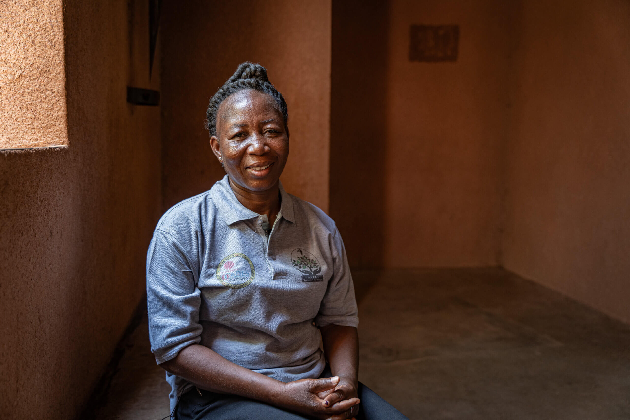 Delphine smiles softly and looks at the camera. Light shines on her through a window, and she sits on a chair in a sand-coloured room. She is wearing dark trousers, a grey polo shirt with a logo of a tree that says “A.V.E.C 1” and an “OCADES Tenkodogo” logo. She has dark hair, which she wears in thick braids in a bun on her head.