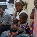 There are five people sitting on a bench, with medical equipment and cables in the background. An adult and child are closest to the camera and out of focus. Delphine looks at a young boy, who has a bandage over his eye, after cataract surgery. He looks relaxed, and sits closely next to his mother, playing with his hands.