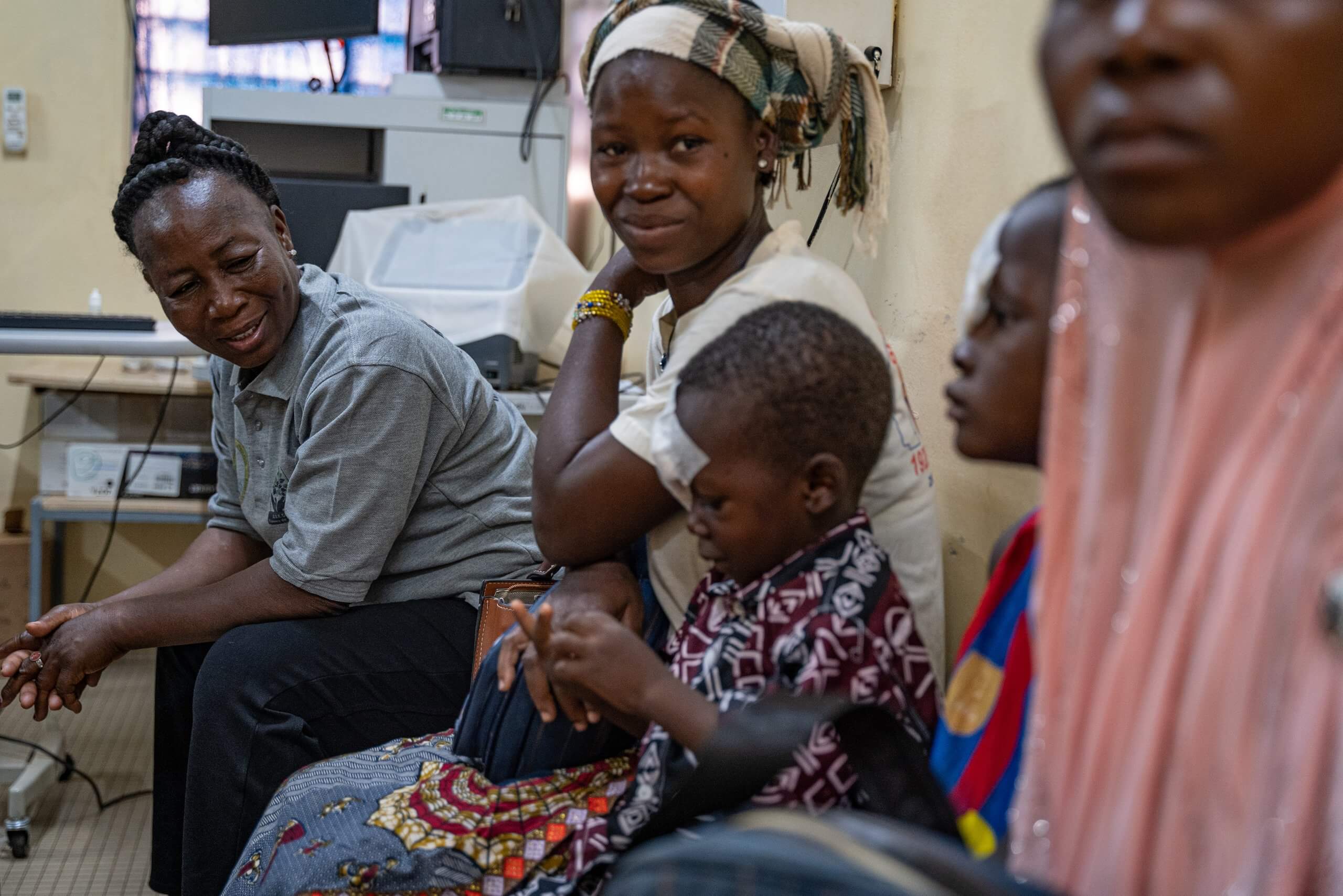 There are five people sitting on a bench, with medical equipment and cables in the background. An adult and child are closest to the camera and out of focus. Delphine looks at a young boy, who has a bandage over his eye, after cataract surgery. He looks relaxed, and sits closely next to his mother, playing with his hands.