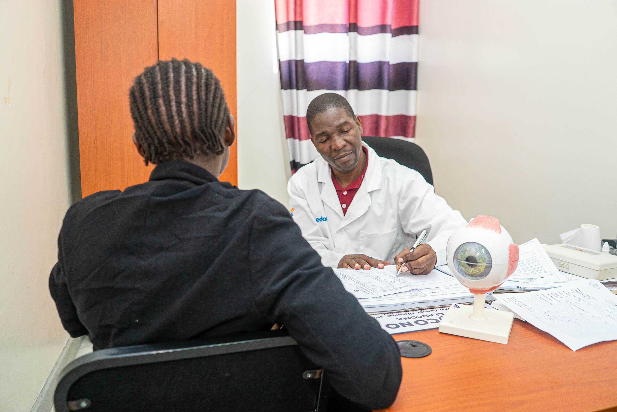 Dr Tiago Machissa sits at a desk writing notes, facing a patient with braided hair seen from behind. A model eyeball and glaucoma information poster sit on the desk between them.