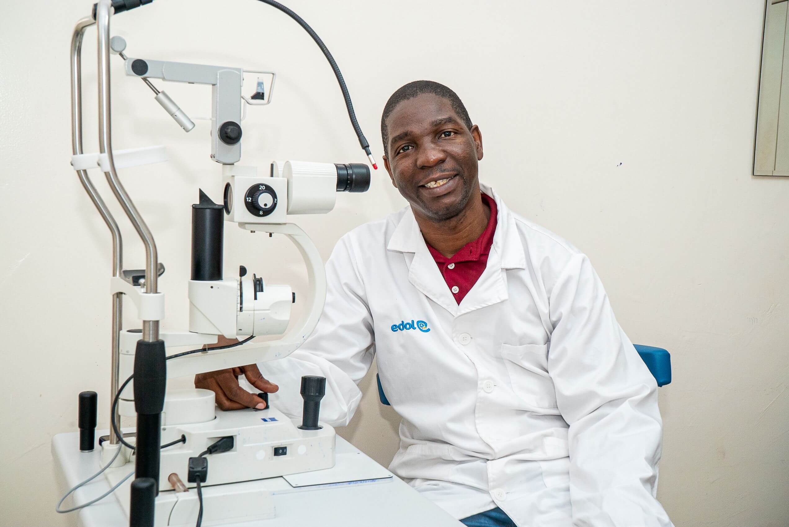Dr Tiago Machissa, a man in a white coat over a maroon polo shirt, sits smiling beside a slit lamp examination machine in a clinic room.