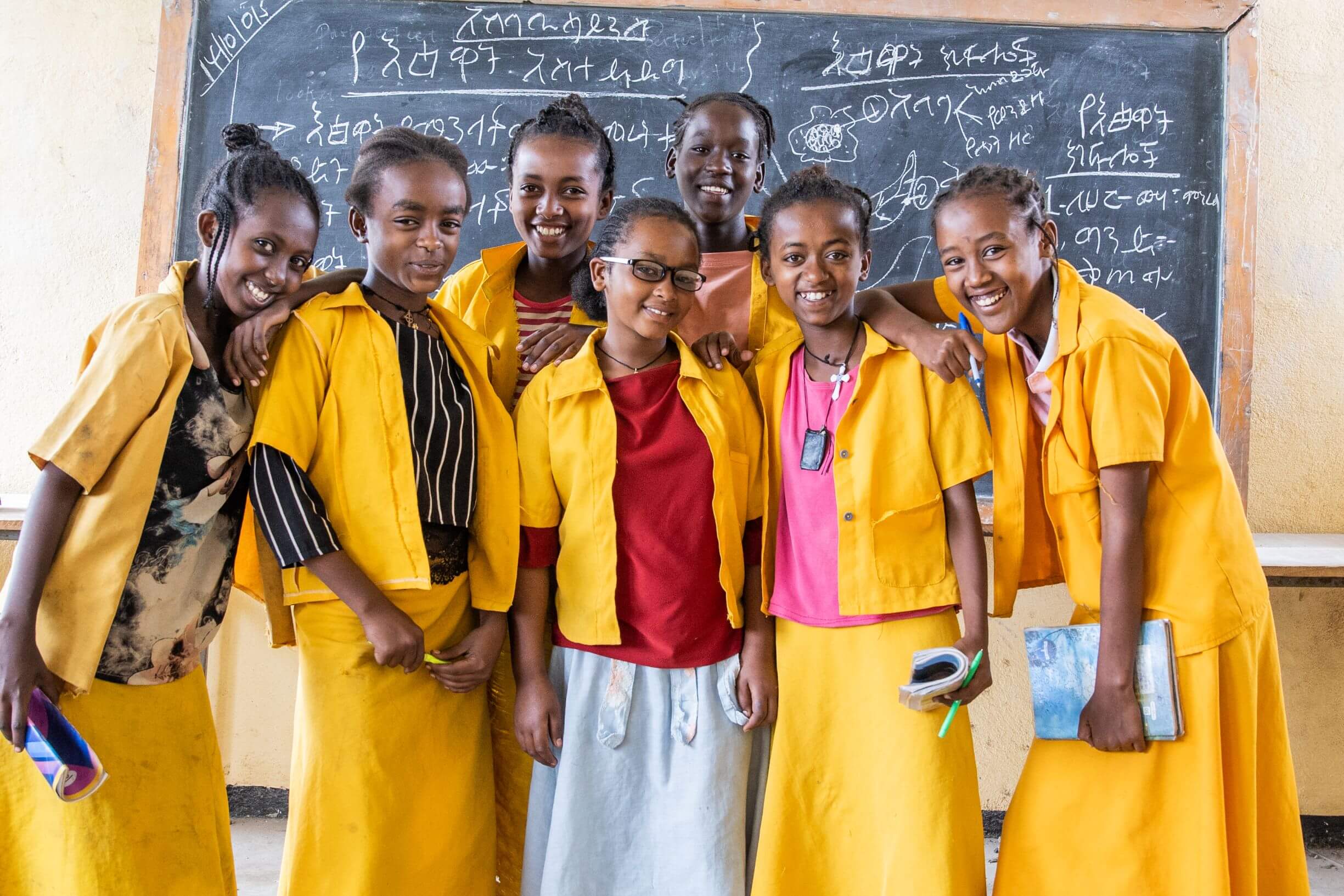 Seven girls in yellow school uniforms, including Nigest, stand together smiling with arms around each other's shoulders in front of a blackboard covered in Amharic writing.