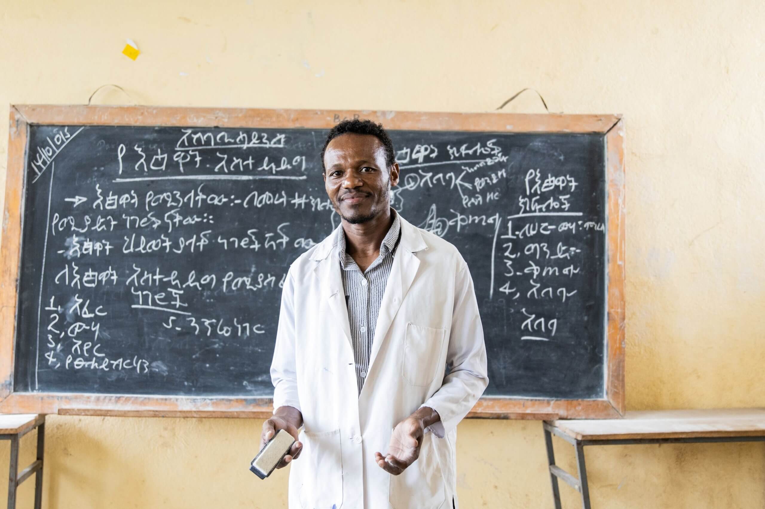 Desalegn Wada, wearing a white coat, stands smiling in front of a blackboard covered in Amharic handwriting, holding a chalkboard eraser.