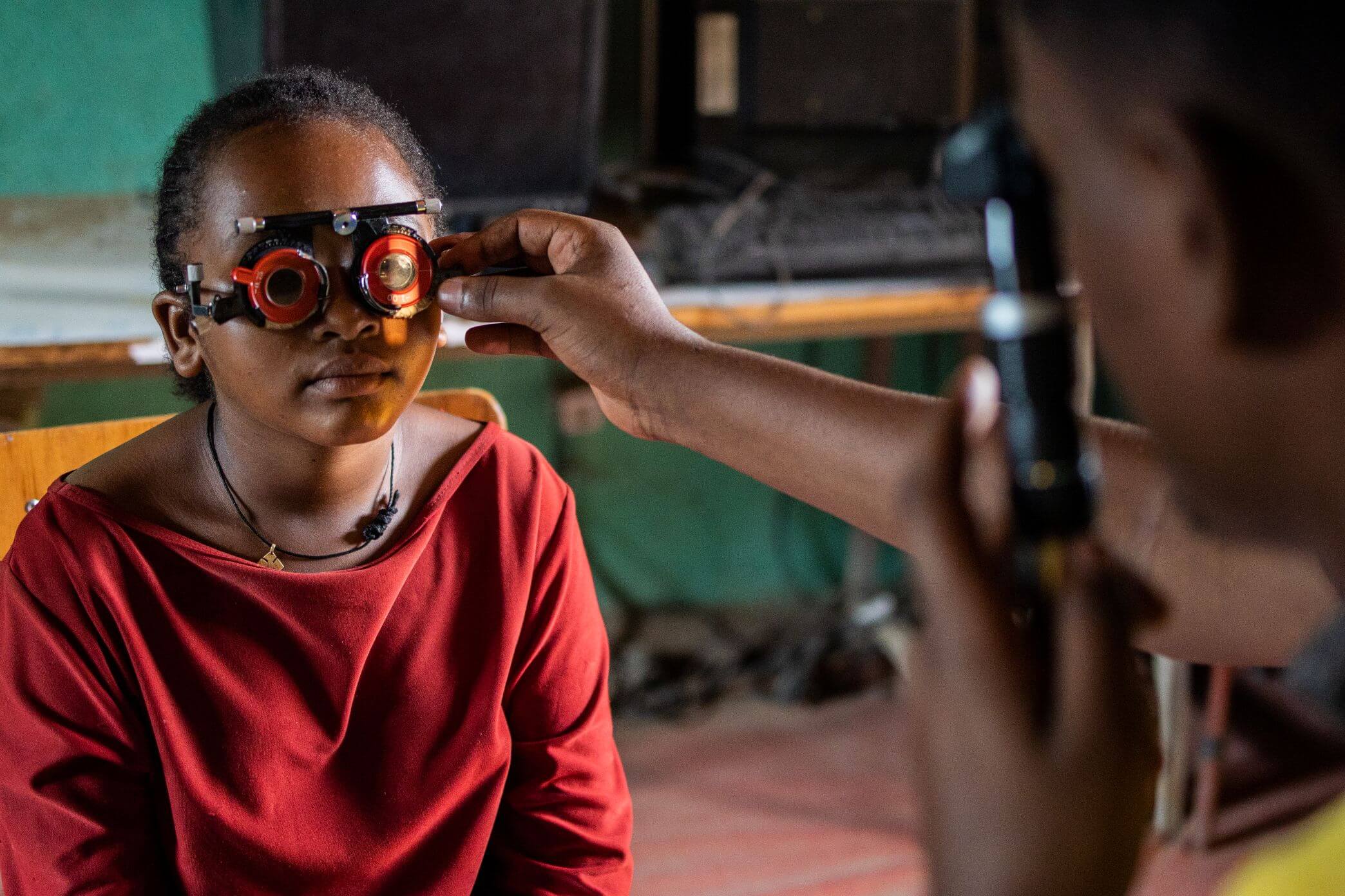 Nigest Unche, wearing a red top, sits wearing a trial frame with a red lens over one eye while a hand holds up a second lens in front of her other eye, against a teal-painted wall.