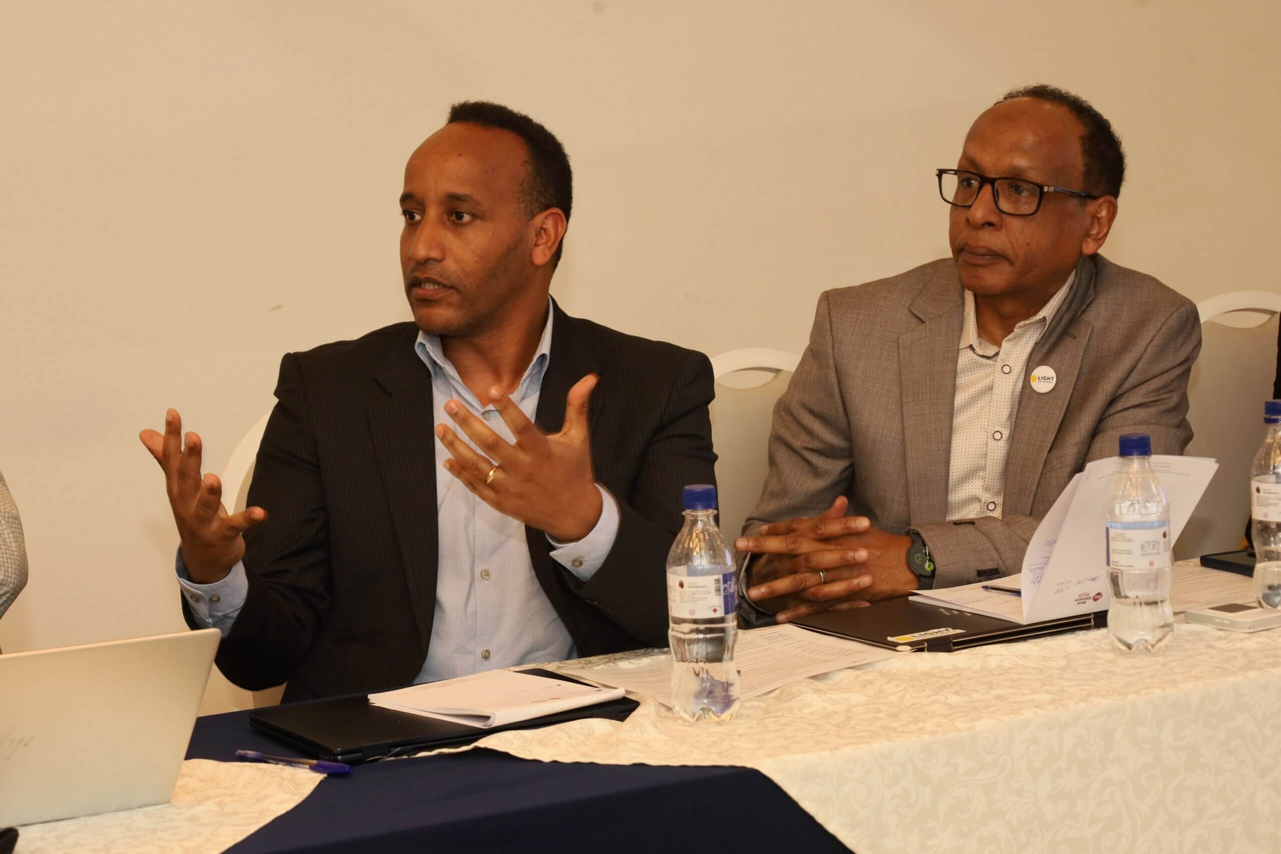 Melaku Tekle (Left), Head of Programmes at Light for the World Ethiopia, and Suadik Hassen, Country Director, sit side by side at a panel table during the media roundtable on eye health in Ethiopia. Melaku gestures as he speaks; Suadik, wearing a Light for the World badge, listens attentively. Water bottles, notebooks and a laptop are on the table in front of them.