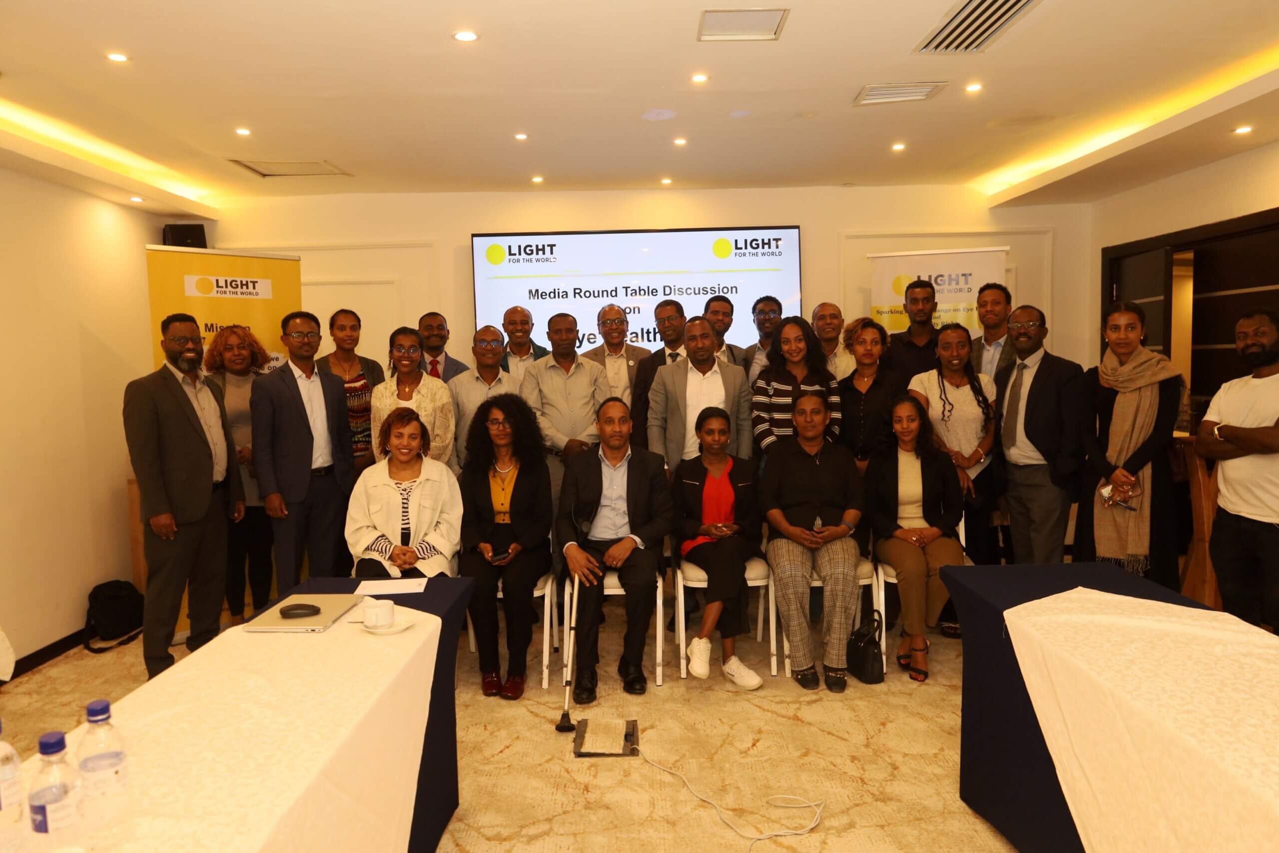 Around 30 participants at Light for the World Ethiopia's media roundtable on eye health pose for a group photo. Behind them, a large screen displays the words "Media Round Table Discussion on Eye Health." Light for the World banners are visible on either side of the room.