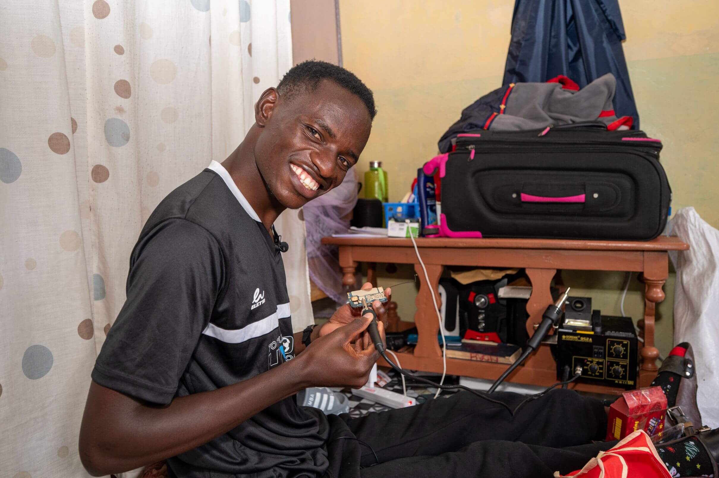 Noah Matara, a young man in a black sports T-shirt smiles at the camera while soldering a small phone circuit board he holds in one hand. He sits indoors near patterned curtains, with a suitcase and soldering equipment on a table beside him.