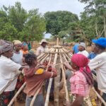 A group of around 15 community members work together to construct the circular wooden roof frame of an accessible storage barn in Mozambique as part of the SPARK programme. Men and women stand around the structure, hands on the timber frame, in a rural outdoor setting.