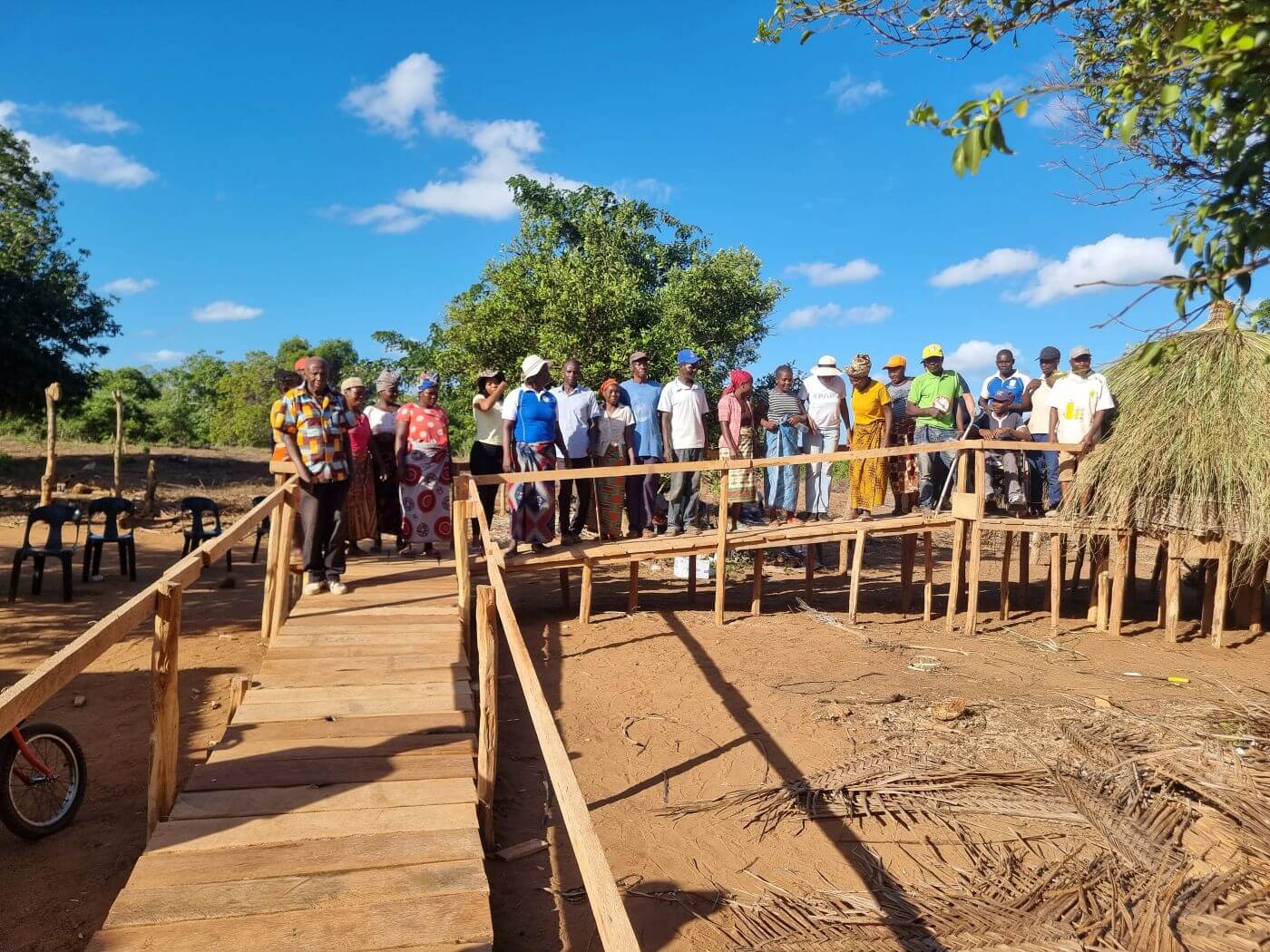 A large group of community members stand together behind a newly completed wooden wheelchair-accessible ramp leading to a storage barn, built as part of the SPARK programme in Mozambique. One participant uses a wheelchair. Blue sky and trees are visible in the background.