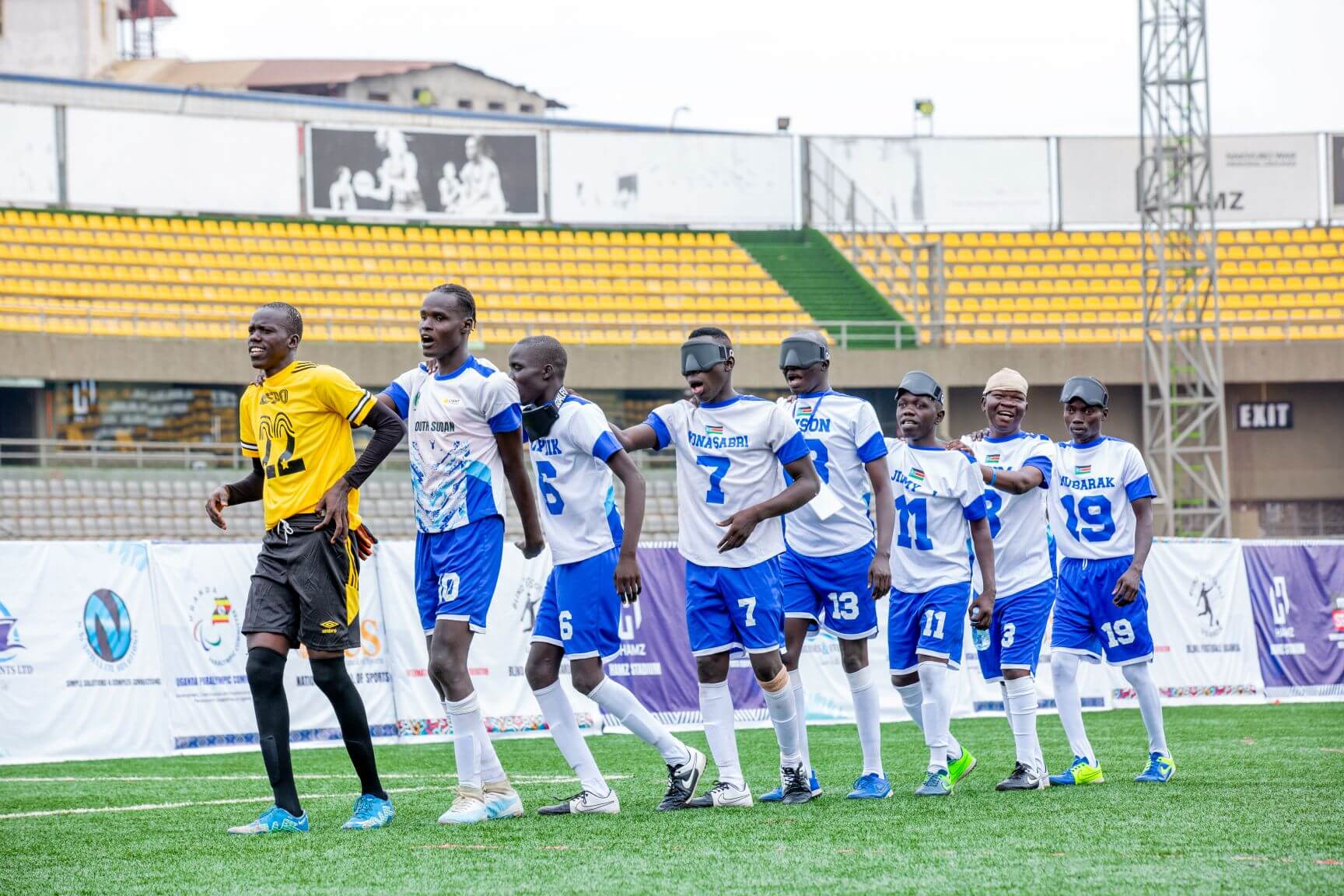 Six South Sudan blind football players, wearing white and blue South Sudan kits and eyeshades, celebrate together on the pitch after winning the 2025 African Blind Football Championship - Division 2 in Kampala, Uganda.  Yellow stadium seating and competition banners are visible in the background.